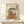 May include: Framed illustration of a charming two-story Japanese bakery with blue tile roof, wooden storefront displaying baked goods, small balcony with plants, round window, bicycle parked outside, and overhead power lines. The artwork has soft beige tones and sits on a light wood shelf next to a small white wrapped gift against a cream wall.