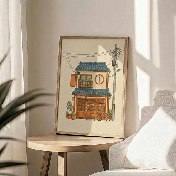 May include: Framed illustration of a two-story Japanese building with blue tile roof, wooden storefront, circular window, small balcony with plants, bicycle parked outside, and power lines overhead. Artwork sits on round wooden side table next to beige armchair with white pillow, green plant leaves visible in corner, natural sunlight casting shadows on cream wall.