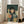 May include: Framed artwork leaning against white wall next to beige sofa, showing illustration of young girl with black bob haircut wearing polka dot dress, surrounded by multiple cats in various colors including orange, gray, blue, and black, set against dark green rainy background with vertical rain lines, rendered in whimsical children's book art style with soft textures and muted tones