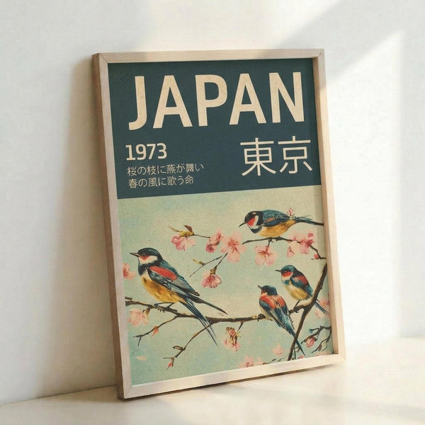 A framed vintage-style poster featuring colorful birds perched on cherry blossom branches. The design has a teal green header with "JAPAN" in cream letters, "1973" and Japanese characters including "東京" (Tokyo). Four small birds with blue, orange and yellow plumage sit among delicate pink sakura flowers against a soft mint green background. The poster is displayed in a light wood frame, leaning against a white wall.