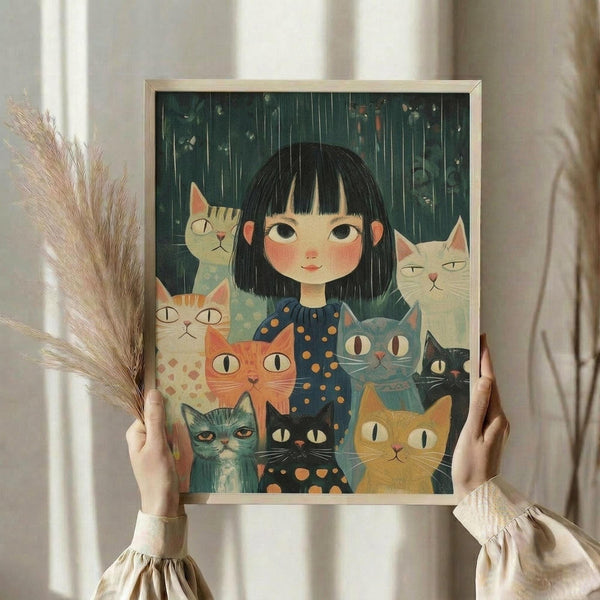 May include: Person holding framed artwork showing young girl with black bob haircut surrounded by multiple cats in various colors including orange, white, gray, and black, set against dark green rainy background. Frame appears light wood tone. Person wears cream colored ruffled sleeve top and holds dried pampas grass stems. Soft natural lighting against neutral curtained background.