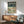 May include: vintage-style travel poster featuring colorful birds perched on cherry blossom branches against mint green background, displayed in wooden frame above mid-century modern walnut credenza with white ceramic lamp, beside teal upholstered armchair with wood arms in bright living room with white walls and natural sunlight