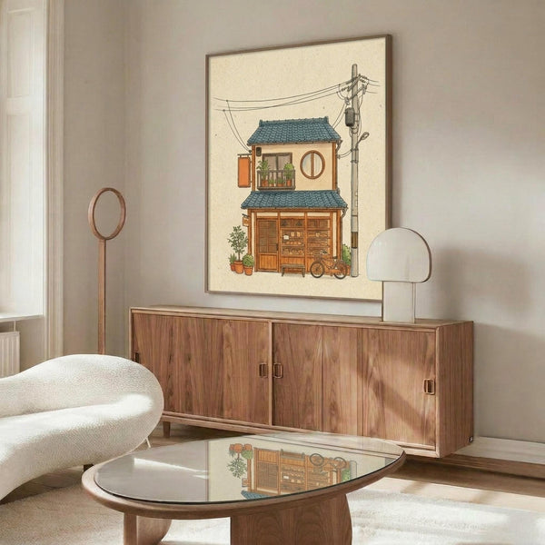 May include: Framed illustration of a cozy two-story Japanese building with blue tile roof, wooden storefront, circular window, small balcony with plants, and power lines overhead. The artwork sits above a walnut credenza in a minimalist room with white curved chair, round glass coffee table, and modern lamp. The print features warm earth tones and detailed architectural elements in a charming street scene style.