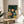 May include: Framed poster print displayed on round wooden side table next to beige upholstered chair. Artwork shows young girl with black bob haircut and polka dot dress surrounded by multiple colorful cats in various shades of orange, blue, and gray against teal background with rain. Natural lighting creates soft shadows across white wall. Plant leaves visible in corner.