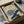 May include: Japanese woodblock print artwork in black frame showing two chrysanthemum flowers - one cream colored, one blue-green - with dark blue leaves and Japanese text. Print is packaged in cardboard box with tissue paper and wooden dowels, photographed on white fabric surface next to dark glass object.