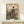 May include: Japanese woodblock print artwork in wooden frame showing woman in ornate purple and floral kimono standing by wooden gate, with cherry blossom trees and mountains in background, displayed on light wood shelf next to small white wrapped gift against cream wall