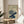 May include: Japanese botanical artwork print in wooden frame showing two chrysanthemum flowers - one purple with blue-green petals and one cream-colored bloom above it, with dark blue-green leaves and stems. Traditional calligraphy text appears on left side. Print displayed on round wooden side table next to beige upholstered chair with white throw pillow, plant leaves visible in background, soft natural lighting.