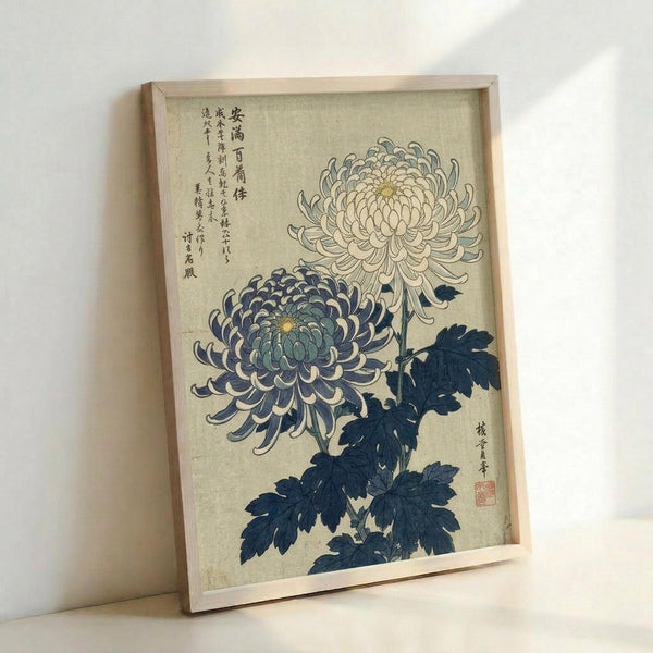 May include: Framed vintage Japanese botanical print showing two chrysanthemum flowers - one blue-purple and one cream-colored - with dark indigo leaves and stems. Features traditional Japanese calligraphy text on aged beige paper background. Displayed in light wood frame leaning against white wall, casting soft shadows.