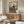 May include: Japanese woodblock print style artwork displayed above wooden credenza in modern living room. Print features woman in colorful traditional kimono with purple, red and blue patterns standing by garden gate, cherry blossoms and mountains in background. White sculptural chair and glass coffee table with wood base visible. White table lamp on credenza.