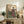 May include: Japanese woodblock print artwork in light wood frame showing woman in colorful kimono standing by wooden gate with cherry blossoms, displayed on rustic wooden sideboard alongside books, ceramic vase, and fern plant in neutral-toned living room setting with beige chair visible.