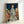 May include: Framed botanical portrait artwork leaning against white wall showing red-haired woman in profile wearing blue and cream leaf-patterned dress, surrounded by large orange and cream magnolia flowers against deep teal background, displayed in light wood frame