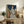 May include: Framed artwork displaying a woman with red hair in profile view wearing a blue and white leaf-patterned dress, surrounded by large brown and cream magnolia flowers against a deep teal background. The painting sits on a rustic wooden sideboard alongside books, a ceramic vase, and a fern plant in a bright, naturally-lit living room with neutral walls and a beige armchair.