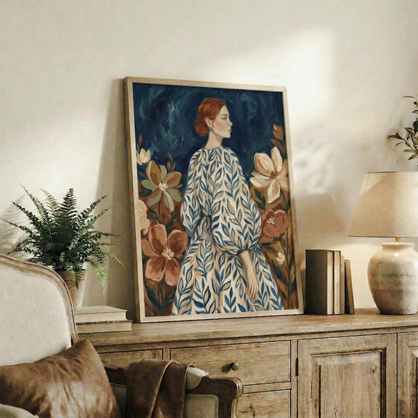 May include: Framed artwork displaying a woman with red hair in profile view wearing a blue and white leaf-patterned dress, surrounded by large brown and cream magnolia flowers against a deep teal background. The painting sits on a rustic wooden sideboard alongside books, a ceramic vase, and a fern plant in a bright, naturally-lit living room with neutral walls and a beige armchair.