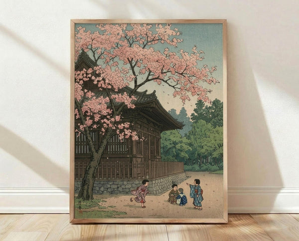 May include: Framed vintage Japanese woodblock print showing a traditional temple building with curved rooflines beside a cherry blossom tree with pink flowers. Three small figures in colorful kimonos play in the courtyard below. The artwork features muted colors with sage green sky, dark wooden architecture, and lush green foliage. Natural wood frame displayed on light wooden floor against white wall with soft shadows.