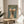 May include: Framed poster featuring an Italian Greyhound wearing a cream knit sweater with colorful pom-pom ear warmers against a sage green background, with "DOGUE" in gold lettering at top. Poster sits on round wooden side table next to beige upholstered chair with white pillow, green plant leaves visible in soft natural lighting.