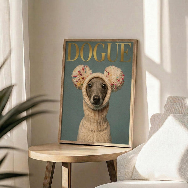 May include: Framed poster featuring an Italian Greyhound wearing a cream knit sweater with colorful pom-pom ear warmers against a sage green background, with "DOGUE" in gold lettering at top. Poster sits on round wooden side table next to beige upholstered chair with white pillow, green plant leaves visible in soft natural lighting.