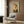 May include: Framed poster featuring a Pomeranian dog wearing olive green military-style jacket and beret against pale background with red "DOGUE" text at top, displayed on wood paneled wall above modern gray upholstered armchair with geometric patterned pillow, alongside gold floor lamp and dark side table with open book