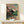 May include: Framed Japanese woodblock print showing a traditional temple building with pink cherry blossoms and people in colorful robes gathered nearby, displayed on a light wood shelf next to a small white wrapped gift with greenery accent against a cream wall.