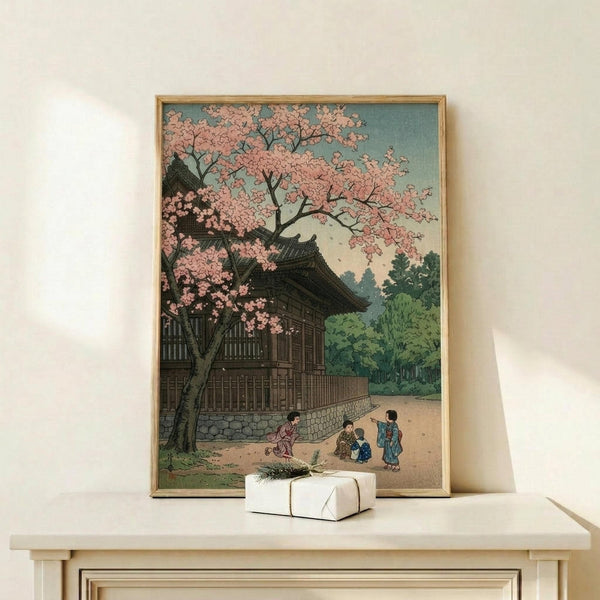 May include: Framed Japanese woodblock print showing a traditional temple building with pink cherry blossoms and people in colorful robes gathered nearby, displayed on a light wood shelf next to a small white wrapped gift with greenery accent against a cream wall.