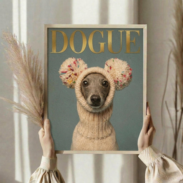 May include: Person holding framed poster featuring Italian greyhound wearing cream knitted sweater with colorful pom-pom ears against sage green background. Gold "DOGUE" text at top mimics fashion magazine style. Light wooden frame, neutral home setting with dried pampas grass and white curtains visible.
