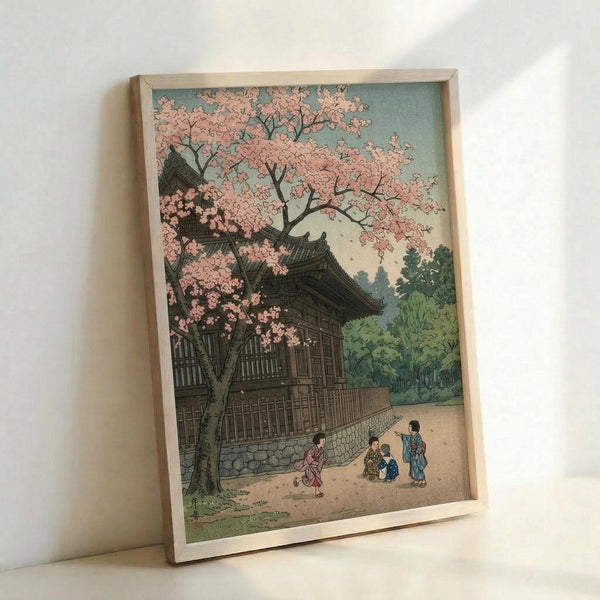 May include: Framed Japanese woodblock print reproduction leaning against white wall. Artwork depicts traditional temple building with curved roofline surrounded by pink cherry blossoms in full bloom. Small figures in colorful kimono walk along stone pathway. Muted color palette of soft pinks, greens, and browns in classic ukiyo-e style. Light wooden frame sits on white surface casting gentle shadow.