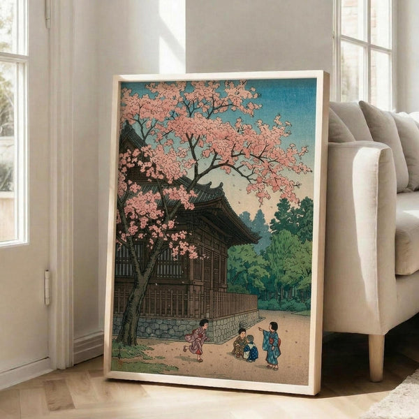 May include: Large framed Japanese woodblock print leaning against white wall next to beige armchair. Artwork shows traditional temple with curved roofline beneath blooming pink cherry blossom tree, with small figures in colorful robes on sandy path. Blue sky background with green foliage. Print has warm wooden frame and displays rich colors in classic ukiyo-e style against bright, sunlit room with windows.