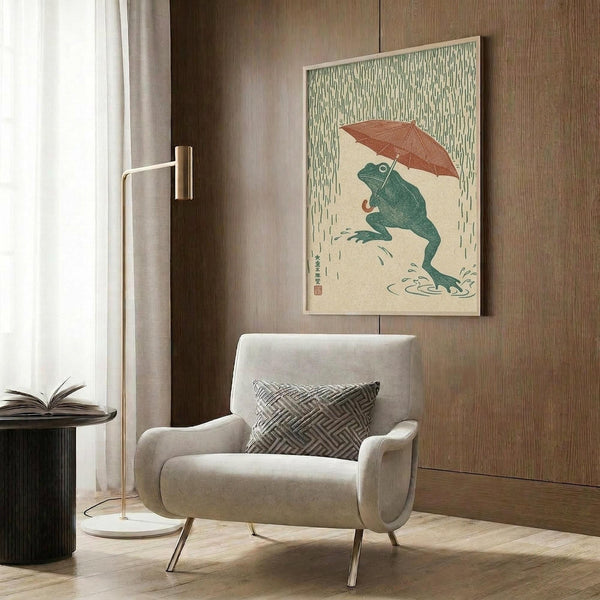 May include: Japanese-style artwork showing a green frog holding a reddish-brown umbrella in falling rain, displayed in a light wooden frame on wood paneled wall. Modern beige armchair with geometric patterned pillow sits below. Brass floor lamp and dark round side table with open book complete the neutral-toned room setting with natural lighting from window.