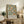 May include: Framed botanical poster featuring orange fruit and green foliage pattern displayed on rustic wooden sideboard beside fern plant, books, and ceramic vases in warm-toned living room setting with beige armchair and white walls