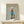 May include: Framed poster print displayed on light wood console table featuring cream-colored poodle wearing turquoise turtleneck sweater against pale blue background. Gold "DOGUE" text at top mimics fashion magazine cover style. Natural wood frame, small white wrapped gift box with twine and greenery sits in front on table surface.