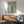 May include: Framed poster featuring a cream-colored poodle wearing a turquoise turtleneck sweater and blue boots, styled as a fashion magazine cover with "DOGUE" in gold lettering. The artwork is displayed above a mid-century modern wooden credenza with a white ceramic table lamp. A sage green upholstered armchair with wooden frame sits nearby. Warm sunlight streams across white walls, creating geometric shadows in this contemporary living room setting.