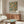May include: Framed botanical poster featuring orange fruits and green foliage with "William Morris" text, displayed above a walnut wood sideboard in a modern living room. White curved chair and glass-top coffee table with blue artwork visible. Warm natural lighting creates shadows across the scene.