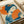Japanese woodblock print style artwork featuring a geisha with dark blue hair adorned with orange flowers, holding an orange folding fan. She wears a blue patterned kimono against a cream background with orange circular elements. The print displays traditional ukiyo-e aesthetics with bold blue and orange colors, visible on white paper with printing materials nearby.