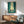 May include: Framed poster displaying two silhouetted samurai warriors dueling with swords in a misty bamboo forest with tall green stalks and golden sunlight filtering through. The artwork has a stylized, vintage-inspired aesthetic with teal and yellow tones. Shown hanging above a mid-century modern wooden credenza with a white ceramic lamp, next to a sage green upholstered armchair with wooden frame in a bright, minimalist living room setting.