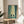 May include: Framed poster showing two samurai warriors dueling with swords in a misty bamboo forest, rendered in green and teal tones with dramatic lighting. The artwork sits on a round wooden side table next to a beige upholstered chair, with a potted plant visible in soft natural lighting from a nearby window.