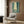 May include: Framed poster showing two samurai warriors dueling with swords in a misty bamboo forest, rendered in blue-green tones with atmospheric lighting. Modern beige armchair with geometric pillow, brass floor lamp, dark round side table with open book, and wood-paneled wall in contemporary living room setting.