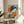 May include: Framed Japanese-style artwork displayed on a round wooden side table next to a beige upholstered chair. The print features a geisha with dark blue hair adorned with orange flowers, holding a bright orange folding fan. She wears a blue patterned kimono against an orange circular background with Japanese characters. The piece is in a thin wooden frame, and soft natural light creates shadows across the neutral-toned room setting with a plant visible in the corner.