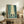 May include: Framed poster print showing two samurai figures dueling with swords in a misty bamboo forest rendered in teal and cream tones, displayed on a rustic wooden sideboard next to a fern plant, ceramic vase, and books in a cozy living room setting with neutral walls and brown leather furniture