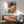 May include: Japanese-style artwork showing a geisha with blue-black hair adorned with orange flowers, holding a vibrant orange fan against an orange background. The poster features Japanese characters in the upper left corner and displays a traditional ukiyo-e art style with bold blues and oranges. Shown framed in light wood hanging above a mid-century modern wooden sideboard with a ceramic table lamp, next to a sage green upholstered armchair in a bright, minimalist living room with white wall