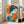 May include: Framed artwork featuring a Japanese woman with blue hair adorned with orange flowers, holding a vibrant orange folding fan. The vintage-style poster uses a blue and orange color palette with Japanese characters in the upper left corner. The piece is displayed in a light wood frame, leaning against a white wall next to a beige upholstered chair in a bright, naturally lit room with hardwood floors.