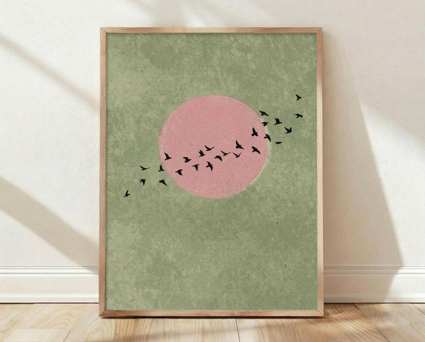 May include: Framed minimalist art print with sage green textured background featuring a large pink circle resembling a sun or moon with black silhouettes of birds flying across and around it. The artwork is displayed in a thin natural wood frame, leaning against a white wall on light wooden flooring with soft natural lighting creating shadows.