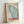 Framed vintage-style travel poster featuring a large blue butterfly wing with delicate shading and detail. Japanese text in red characters runs vertically down the left side, with "The Rising Wind" in cursive script and "JAPAN 1977" and "TOKYO" in bold red letters at the bottom. The poster has a cream background and sits in a light wooden frame, leaning against a white wall with soft natural lighting casting gentle shadows.