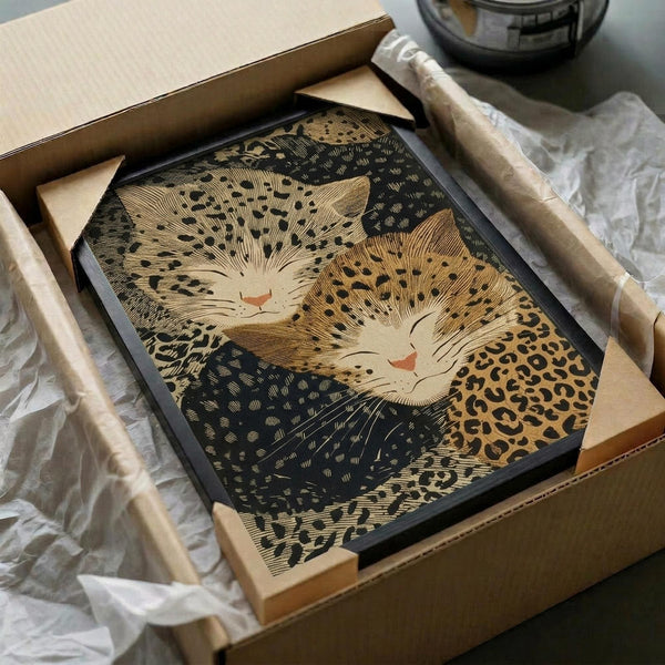 Two sleeping cats curled together in a circular composition, rendered in traditional Japanese woodblock print style with golden-brown and dark tones. The spotted leopard-pattern cats have peaceful expressions with closed eyes and small pink noses. The artwork is displayed in a black frame, nestled in tissue paper within a cardboard shipping box alongside protective packaging materials.