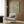 May include: Modern living room with beige armchair featuring geometric patterned pillow, brass floor lamp with white base, dark round side table with open book, and framed wall art showing green textured background with pink circle and silhouetted birds in flight, displayed against warm wood paneled wall with natural lighting from window.