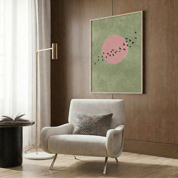 May include: Modern living room with beige armchair featuring geometric patterned pillow, brass floor lamp with white base, dark round side table with open book, and framed wall art showing green textured background with pink circle and silhouetted birds in flight, displayed against warm wood paneled wall with natural lighting from window.