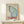 May include: Framed vintage-style poster featuring large blue and teal butterfly wing with Japanese text and red "TOKYO" lettering, displayed on light wood shelf next to small white wrapped gift with greenery sprig, against cream wall with natural lighting