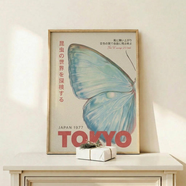 May include: Framed vintage-style poster featuring large blue and teal butterfly wing with Japanese text and red "TOKYO" lettering, displayed on light wood shelf next to small white wrapped gift with greenery sprig, against cream wall with natural lighting