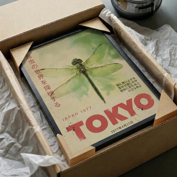 May include: Vintage-style dragonfly poster in black frame nestled in cardboard shipping box with protective tissue paper. Green and cream colored print features detailed dragonfly illustration with Japanese text and "TOKYO" in bold red letters, dated 1977. Wooden dowels for hanging visible alongside frame in rustic packaging setup.