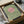 May include: Framed minimalist art print being unboxed from cardboard packaging with tissue paper. Green textured background with coral pink circular sun and black silhouetted birds flying across. Black frame with light wood corners visible in shipping box on white surface.