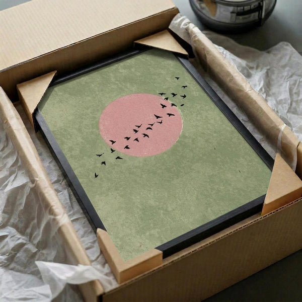 May include: Framed minimalist art print being unboxed from cardboard packaging with tissue paper. Green textured background with coral pink circular sun and black silhouetted birds flying across. Black frame with light wood corners visible in shipping box on white surface.