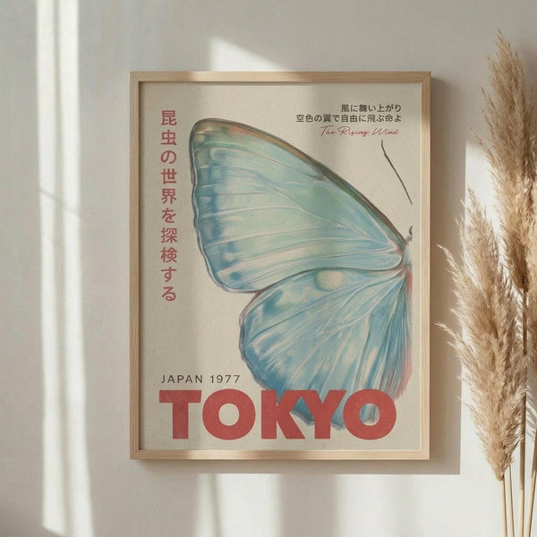 May include: Framed vintage-style poster with blue-green butterfly wing design and Japanese text, mounted on white wall next to dried pampas grass stems. Features "TOKYO" in bold red letters with "JAPAN 1977" above, delicate wing illustration in soft blue and teal watercolor style, housed in light wood frame.